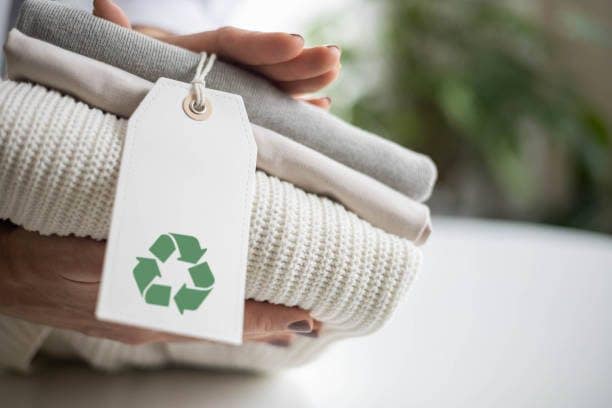Woman's hands hold a stack of clean neat, knitted clothes with a label with a recycling sign. Recycle Icon show on Tag
